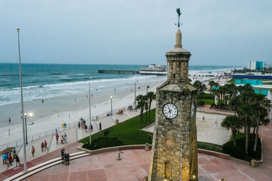 A stone clock tower reading DAYTONA BEACH stands above a beachfront park with palm trees, a pier, and volleyball courts visible nearby.