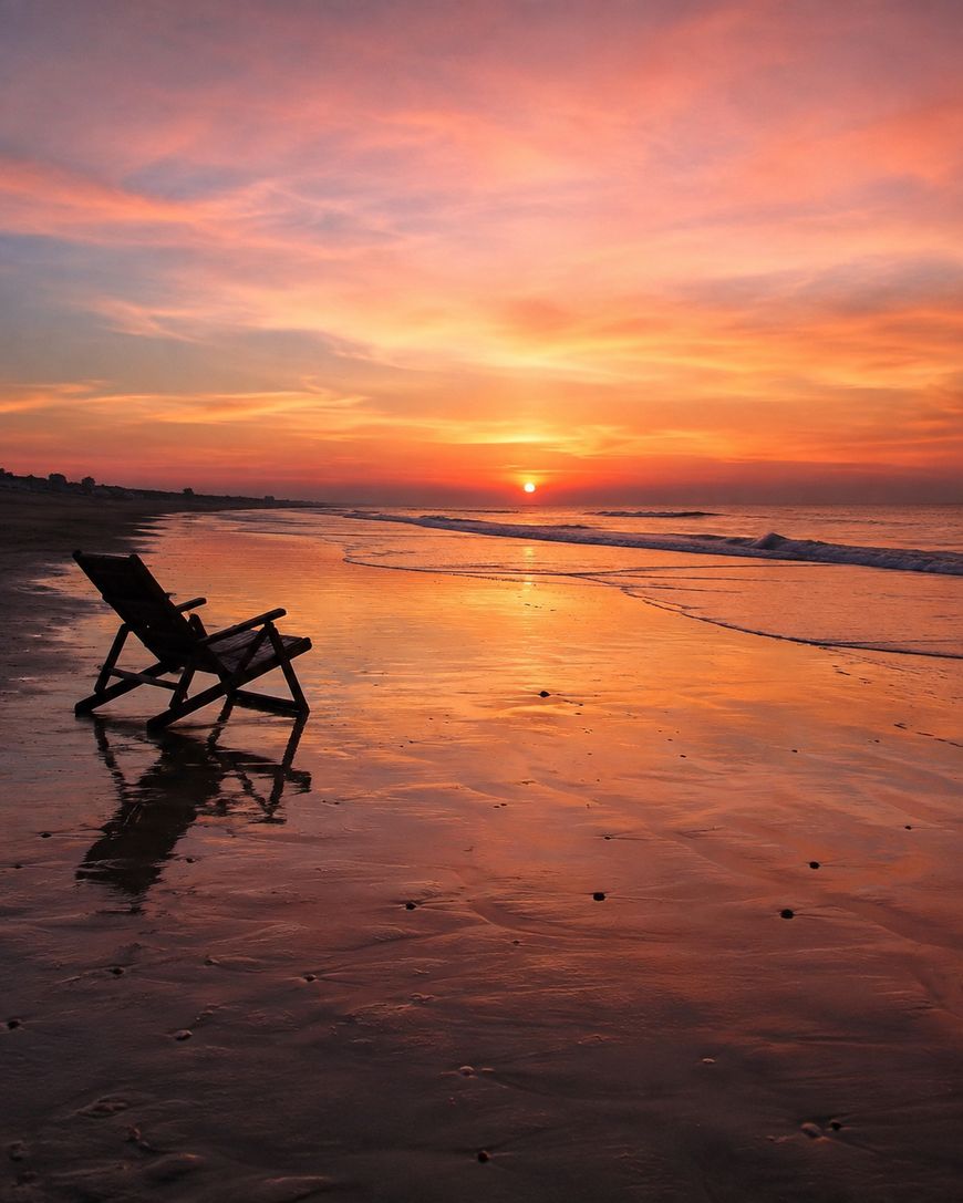 A wooden beach chair faces the ocean at sunset, reflecting vibrant orange, pink, and purple hues on the wet sand. The scene is calm and empty, evoking relaxation and tranquility.
