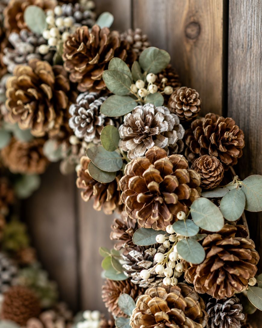 A close-up of a wreath made from pinecones, green eucalyptus leaves, and small white berries, hanging on a rustic wooden wall.