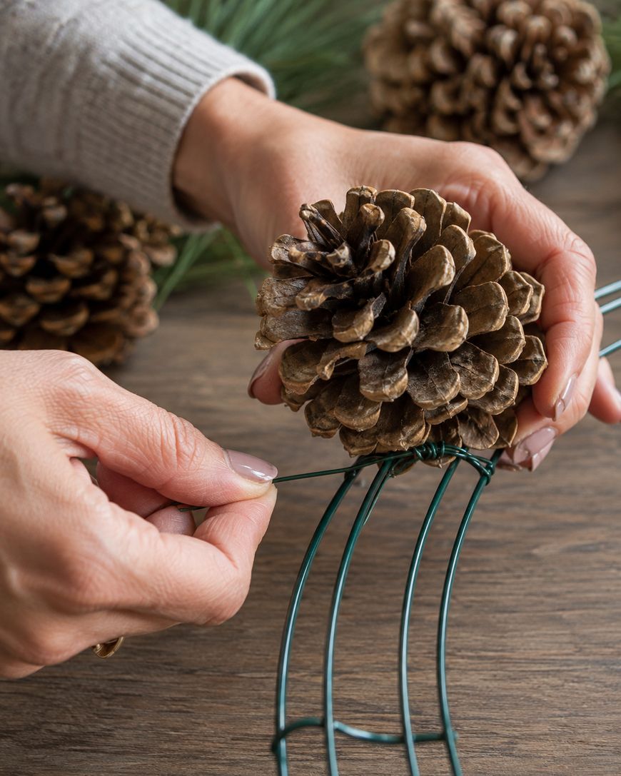 A person attaches a pinecone to a green metal wreath frame using floral wire.