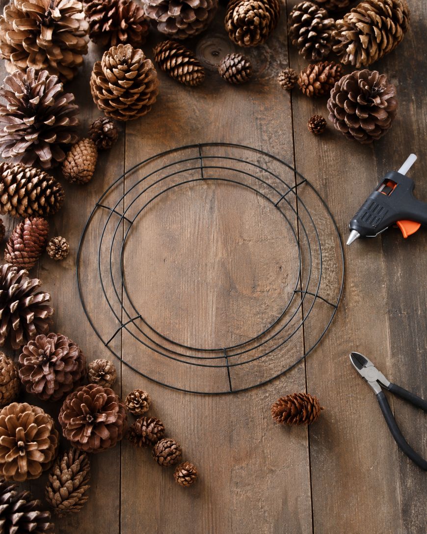 A wire wreath frame, pinecones, hot glue gun, and wire cutters are arranged on a rustic wooden table, ready for a DIY craft project.