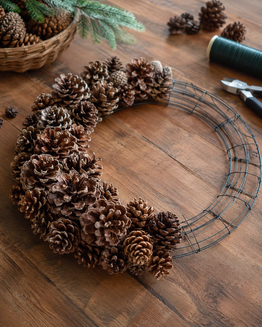 A wire wreath partially covered with pinecones sits on a wooden table with a spool of wire and pliers nearby.