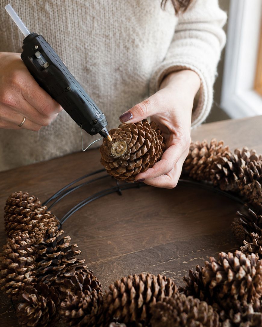 A person uses a hot glue gun to attach a pinecone to a wreath frame on a wooden table.