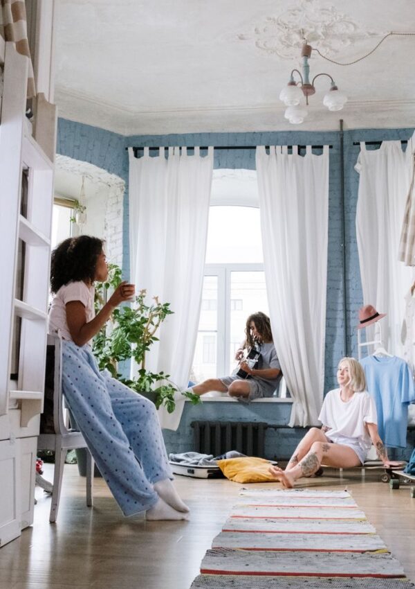 Three women relax in a bright, cozy bedroom with tall windows and white curtains. One sits on a chair with a cup, another on the windowsill, and the third on the floor near a rug and plants.