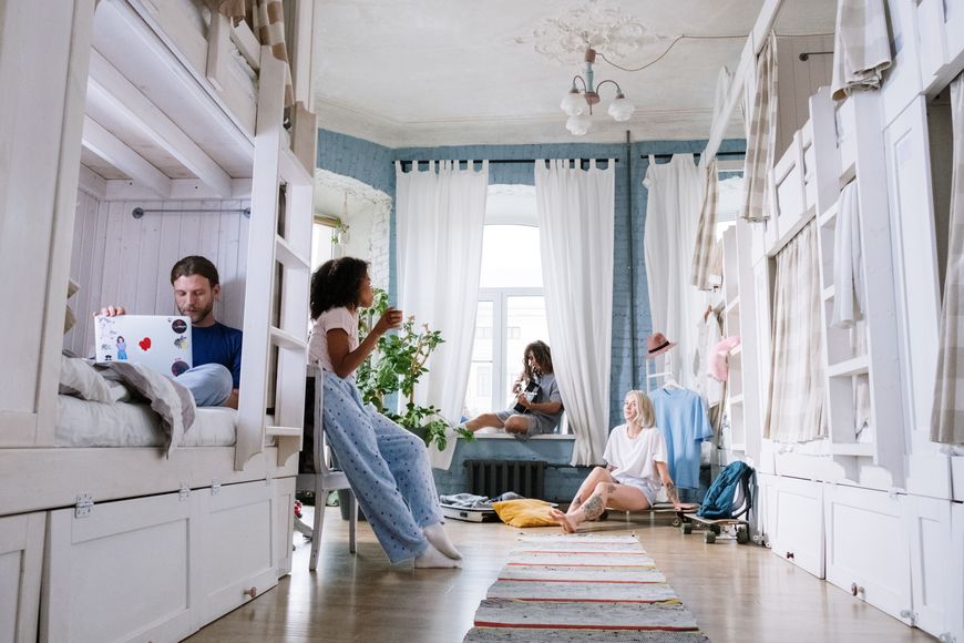 Four people relax in a cozy, bright dorm room with bunk beds, white curtains, and plants. One uses a laptop on a bed, while the others sit and chat on the floor and near the window.