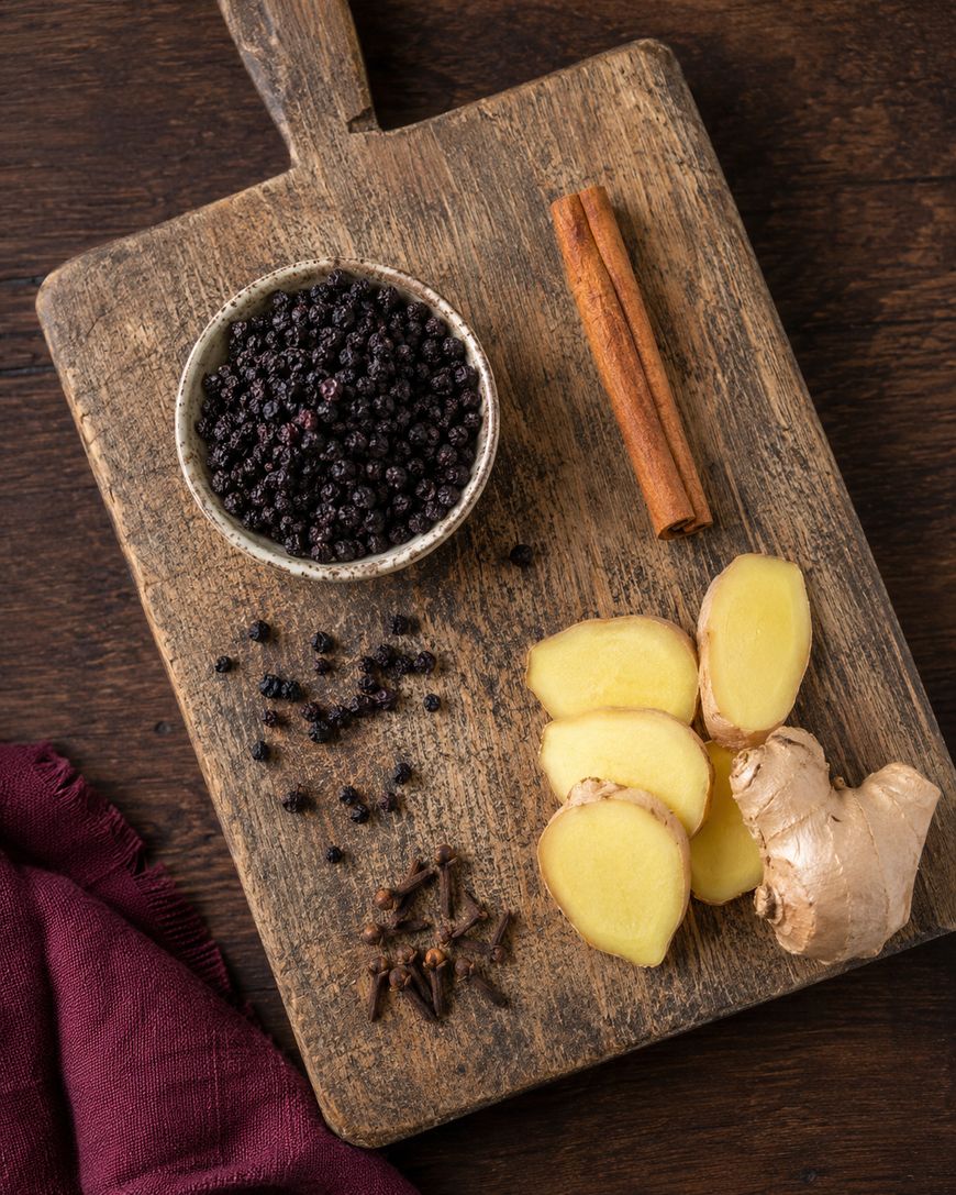 A wooden board with a bowl of elderberries, a cinnamon stick, fresh ginger slices, whole ginger root, whole cloves, and a maroon cloth nearby on a dark wooden table.