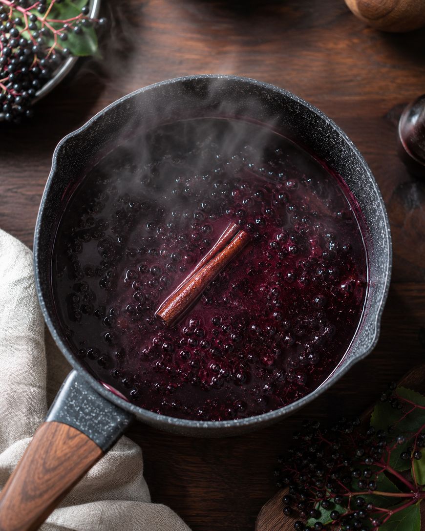 A pot of simmering dark purple liquid with elderberries and a cinnamon stick, releasing steam.
