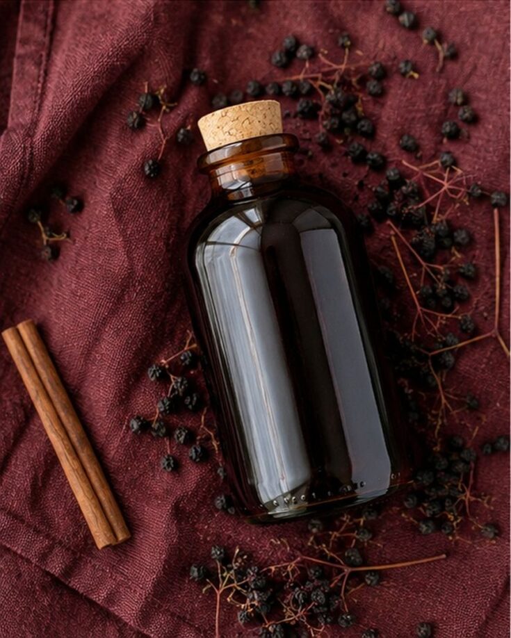 A brown glass bottle with a cork sits on a burgundy cloth, surrounded by dried elderberries and two cinnamon sticks.