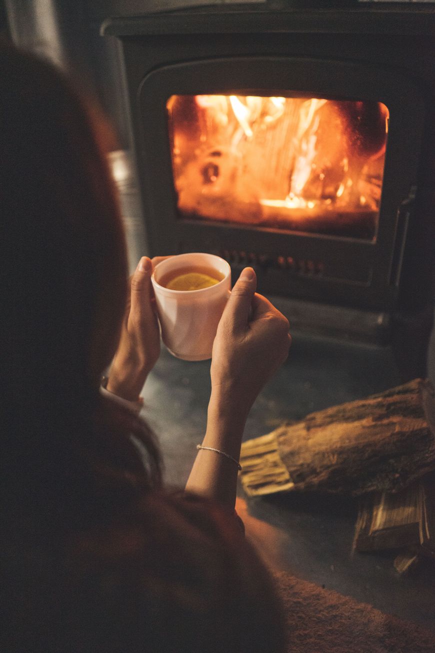 A person holding a mug sits near a wood-burning stove with a fire inside, enjoying their calm evening reset. Logs are on the floor, and the scene feels warm and cozy.