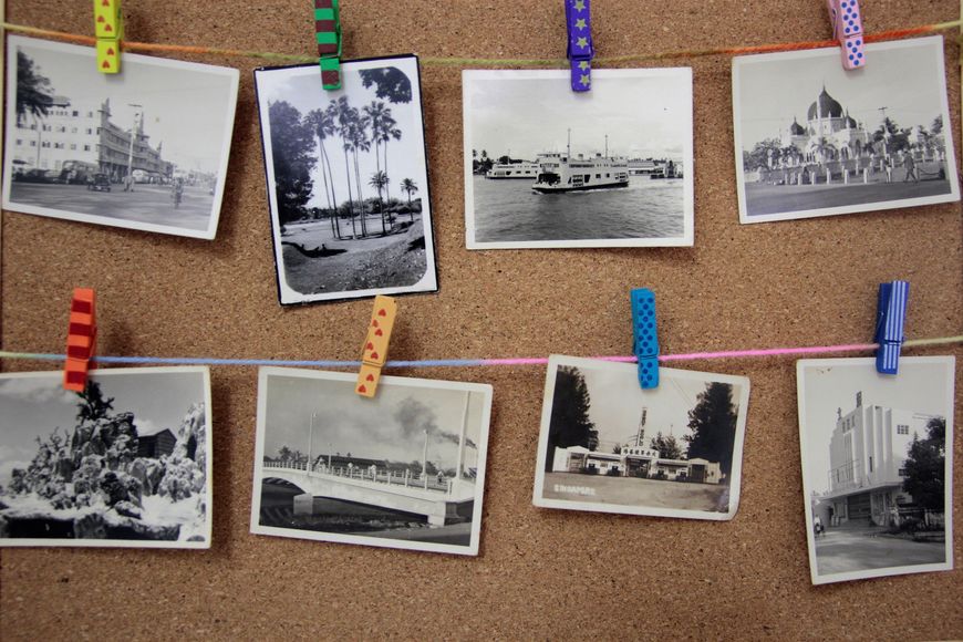 Eight black and white vintage photos are clipped to two strings on a corkboard with colorful clothespins. Each photo depicts various buildings, landmarks, or boats.