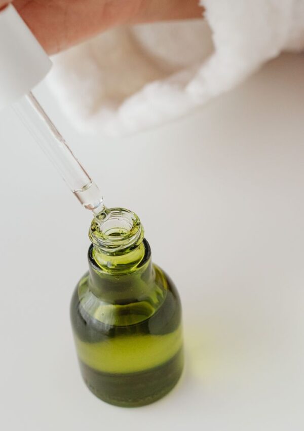 A hand holds a dropper above an open green glass bottle, dispensing a liquid. The background is white and the person appears to be wearing a white robe.