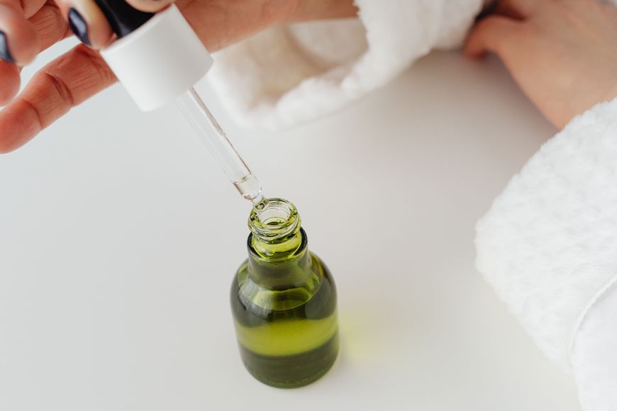 A person in a white robe holds a dropper above a green glass bottle containing liquid, preparing to dispense a drop.