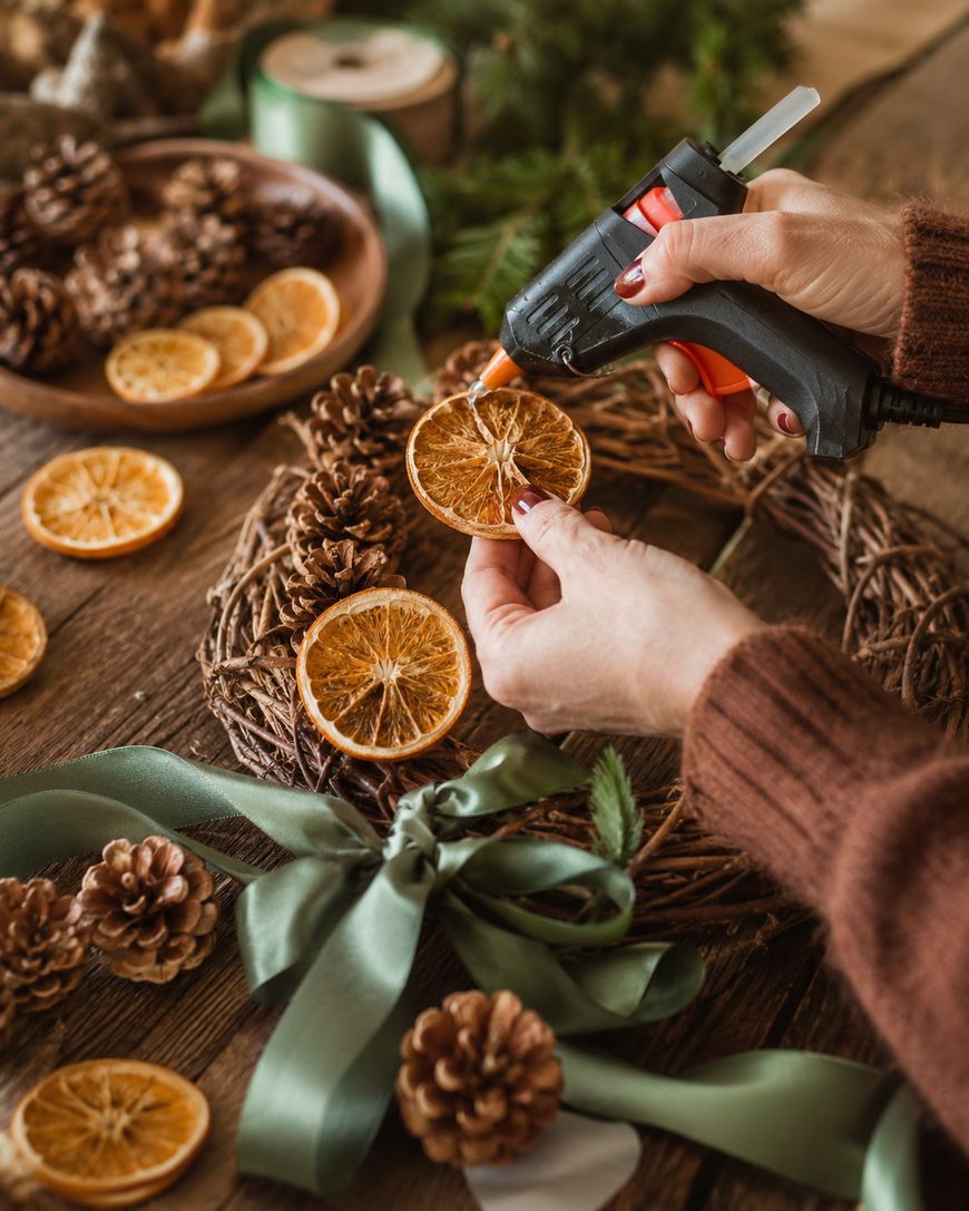 A person uses a hot glue gun to attach dried orange slices to a wreath. Pinecones, ribbon, and craft supplies are on the rustic wooden table.