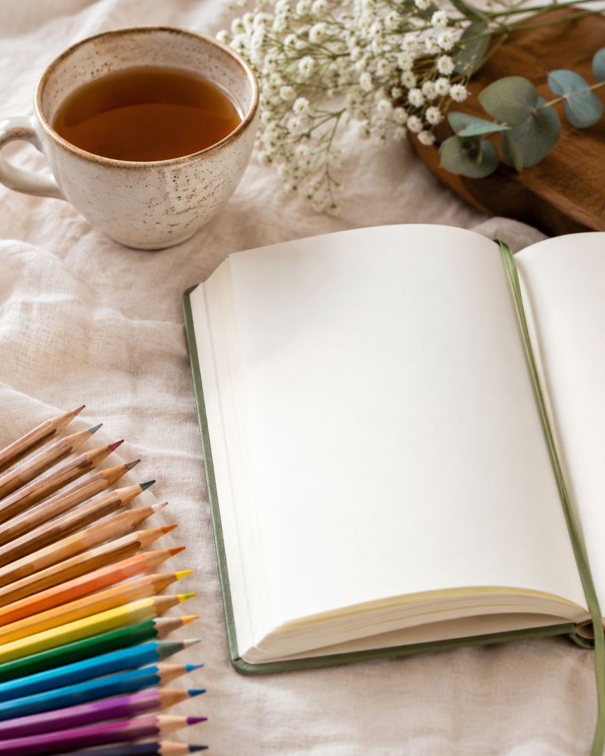 A blank open notebook, colored pencils, a cup of tea, and sprigs of white flowers rest on a soft beige fabric, creating a cozy and creative workspace.