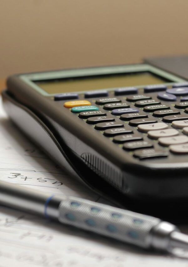 A close-up of a scientific calculator and a pen resting on lined paper with handwritten notes. The background is blurred, focusing on the calculators buttons and the pens grip detail.
