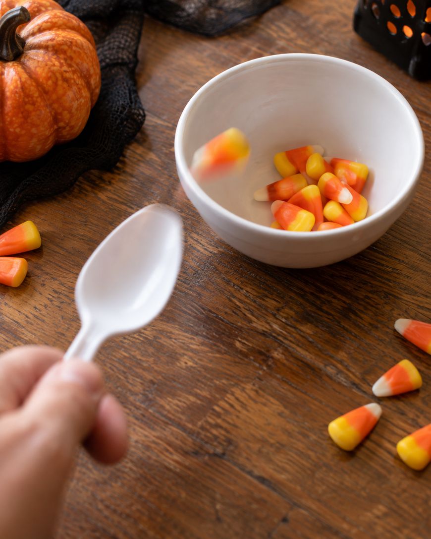 A hand holding a white spoon tosses candy corn into a white bowl on a wooden table. Scattered candy corn and a small pumpkin decorate the scene.
