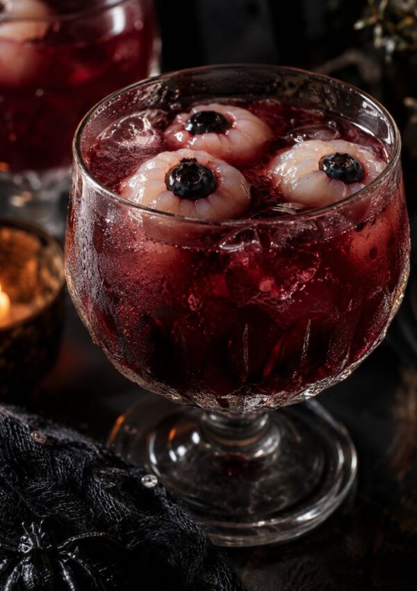 A spooky red cocktail with ice, topped with three lychee eyeballs stuffed with blueberries, served in a glass, with a candle and dark decorations nearby.