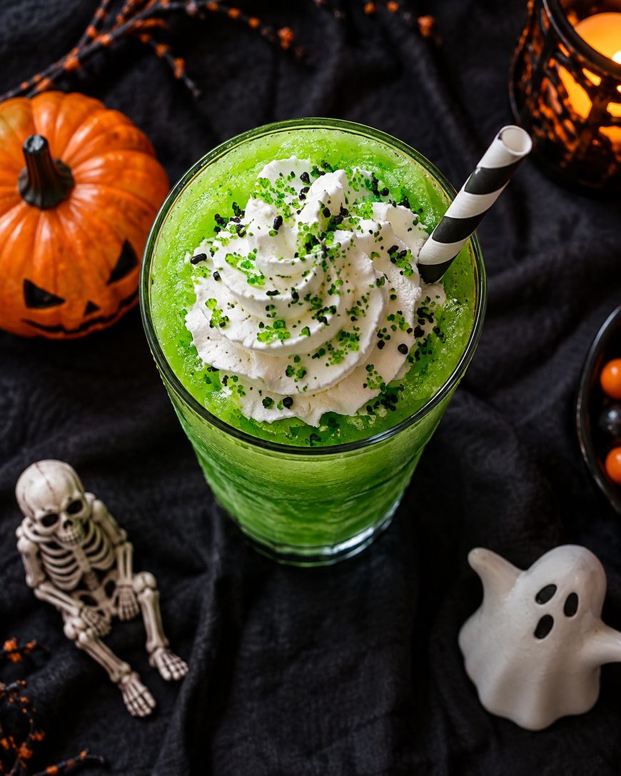 A green slush drink with whipped cream and green sprinkles, served with a striped straw. Halloween decorations, including a pumpkin, skeleton, and ghost figure, surround the glass on a black cloth.