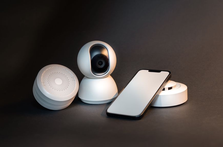 Three smart home devices and a smartphone are arranged on a dark surface. The devices include a security camera, a round speaker, and a round sensor. The phone screen is blank and faces upward.