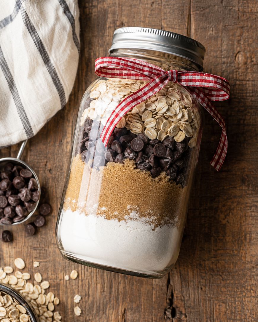 A glass jar filled with layered ingredients—flour, brown sugar, chocolate chips, and oats—topped with a metal lid and a red gingham ribbon, sits on a wooden surface beside a striped cloth.
