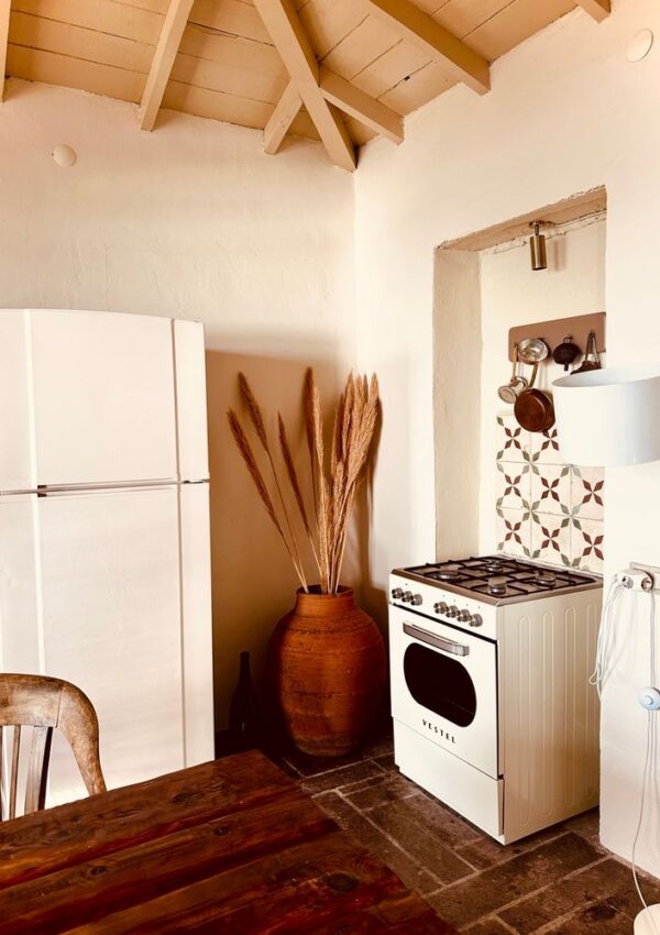 A rustic kitchen with a white fridge, vintage stove, tall ceramic vase with dried grass, wooden table, and patterned backsplash under a light wooden ceiling.