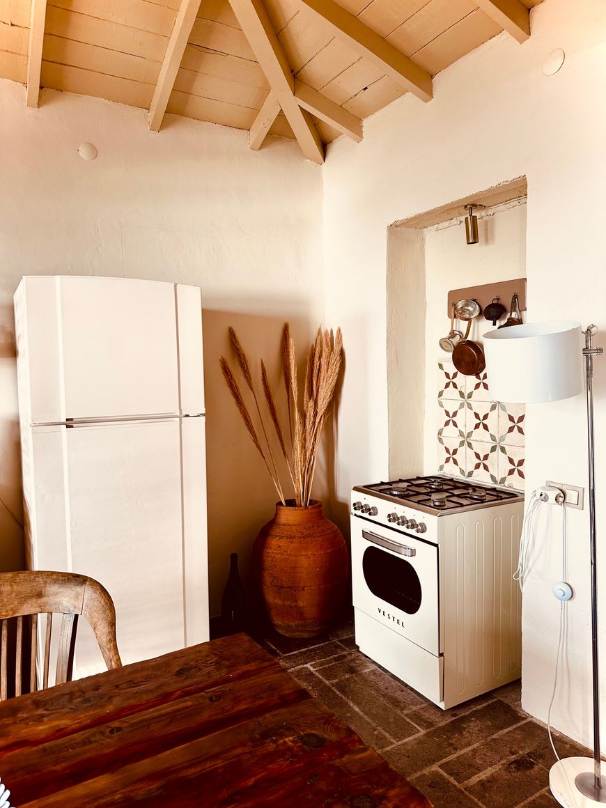 Cozy rustic kitchen with a white fridge, stove, a large clay vase holding dried pampas grass, a wooden table, and tiled backsplash. Exposed wooden ceiling beams add charm.