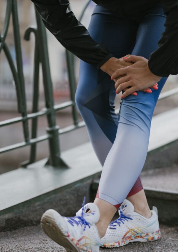 A person in colorful running shoes and blue leggings is standing on stairs, holding their knee as if in pain, near a metal railing outdoors.