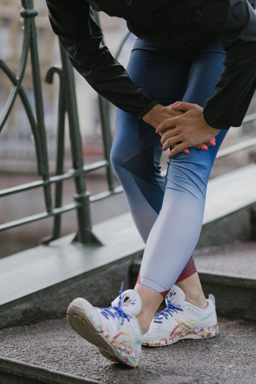 A person in athletic wear holds their knee while standing on outdoor steps, possibly indicating discomfort or injury, with colorful running shoes visible.