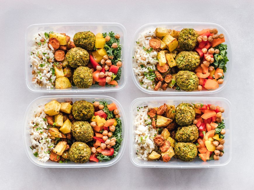 Four plastic containers filled with rice, chickpeas, roasted potatoes, diced carrots, red bell peppers, and green herb falafel are arranged neatly on a light surface—perfect for effortless Meal Prep as you ease into your Fall Schedule or transition to a Back-to-Office routine.