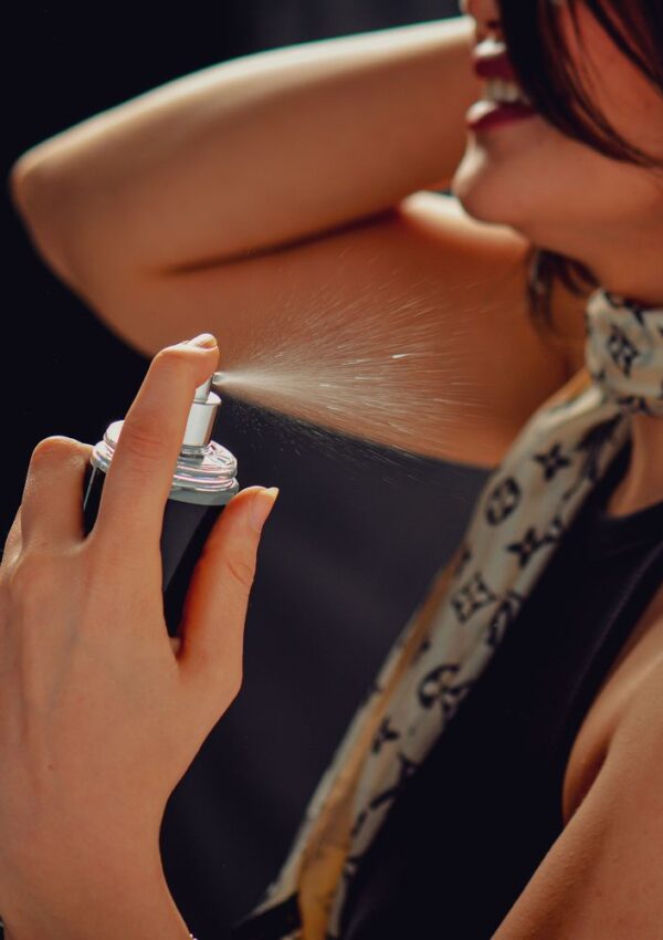 A woman smiles as she sprays perfume onto her neck, holding the bottle close to her skin. She wears a patterned scarf and a dark sleeveless top.