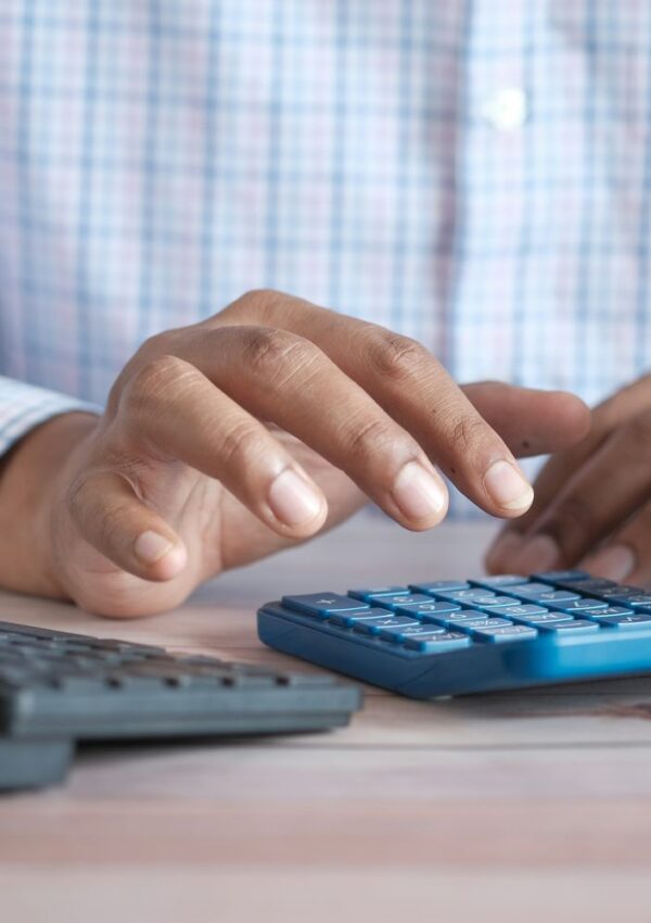 A person in a plaid shirt uses a blue calculator on a wooden desk, with another black calculator nearby.