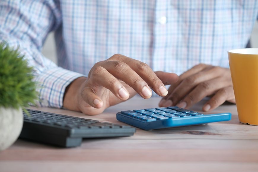 A person in a plaid shirt uses a blue calculator at a desk with a keyboard, yellow cup, and a small potted plant.