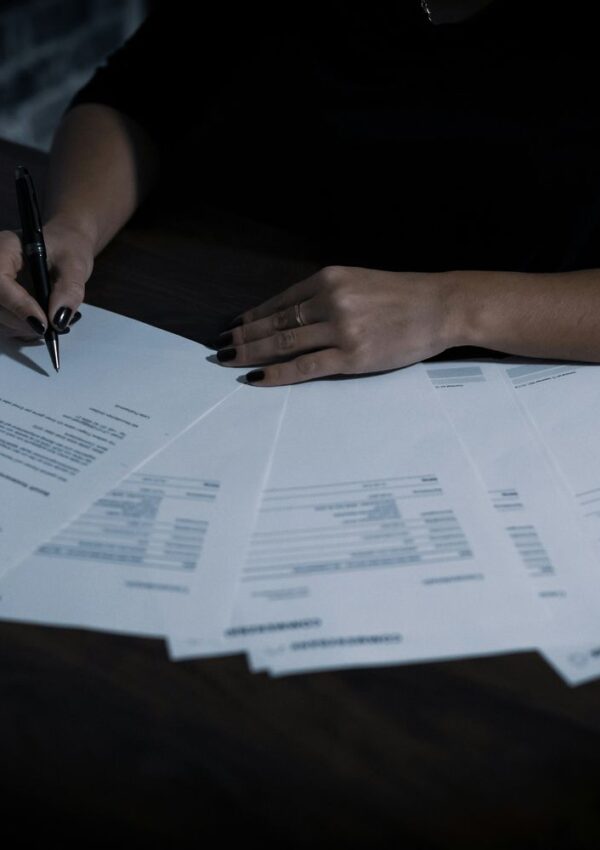 A person with dark nail polish writes on papers spread out on a dark table, appearing to fill out or review documents.