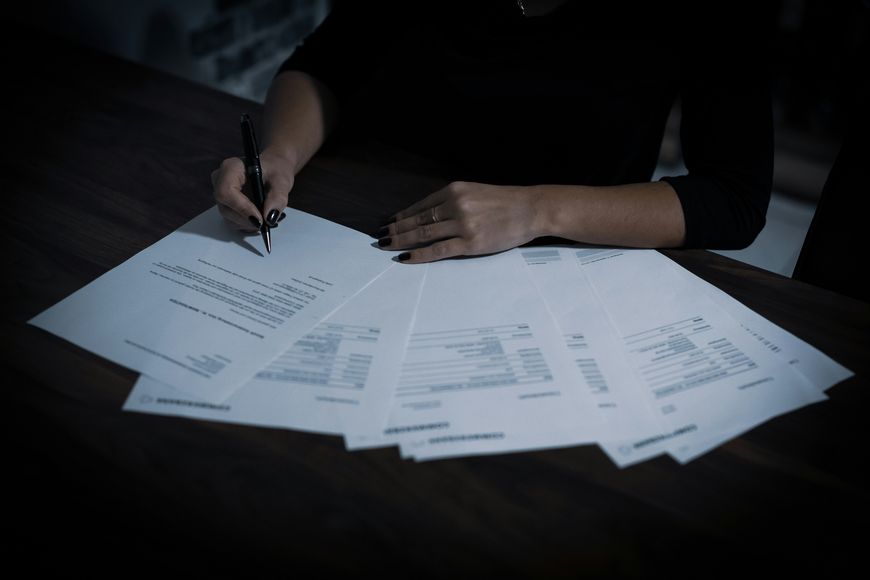 A person in dark clothing signs a document at a desk, surrounded by several sheets of paper with printed text and tables.