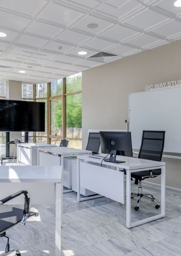 Modern office space with white desks, black chairs, desktop computers, a large TV screen, whiteboard, and large windows letting in natural light.