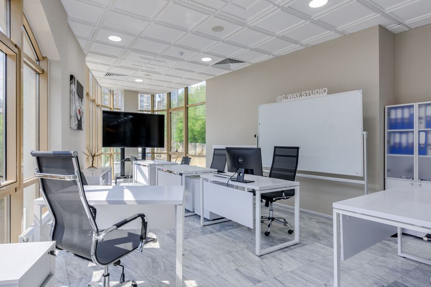 Modern office with white desks and chairs, two computer monitors, a large TV, a whiteboard, and large windows letting in natural light. Marble flooring and minimalistic decor create a clean, bright workspace.