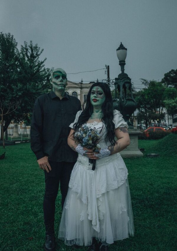 Two people in a park wear skeleton-inspired makeup and dark clothing. One wears a white dress and holds a bouquet, resembling a gothic or Halloween bride. The background has trees, lamps, and historic buildings.