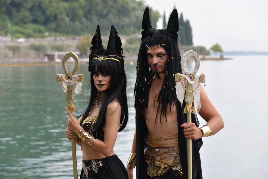 A couple in matching black and gold Egyptian jackal-eared headpieces holding gold ankh staffs by the water.