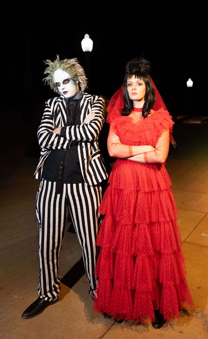 A couple dressed as Beetlejuice and Lydia Deetz, one in a black and white striped suit with white face paint, the other in a red ruffled wedding dress and veil.