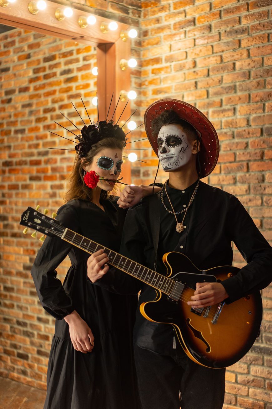 A couple in Day of the Dead sugar skull face paint, one holding a guitar, standing in front of a brick wall with string lights.