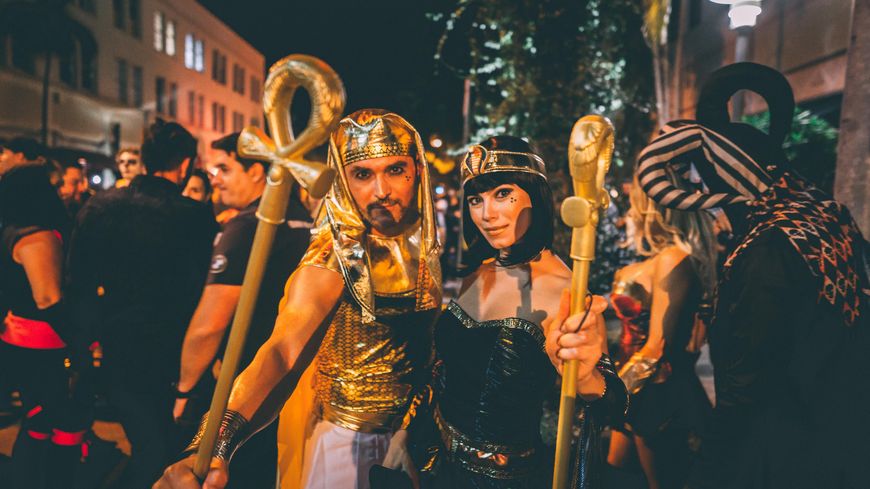 A couple in gold Egyptian pharaoh and queen costumes holding gold staffs at a nighttime street party.