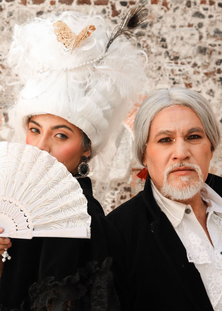 A couple in 18th century aristocrat costumes, one in a tall white powdered wig, the other with gray hair and a black velvet coat.
