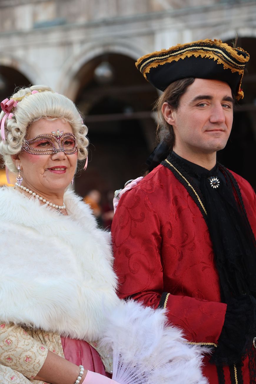 An older couple in elegant masquerade costumes, one in a white wig and jeweled mask, the other in a red and black tricorn hat and coat.