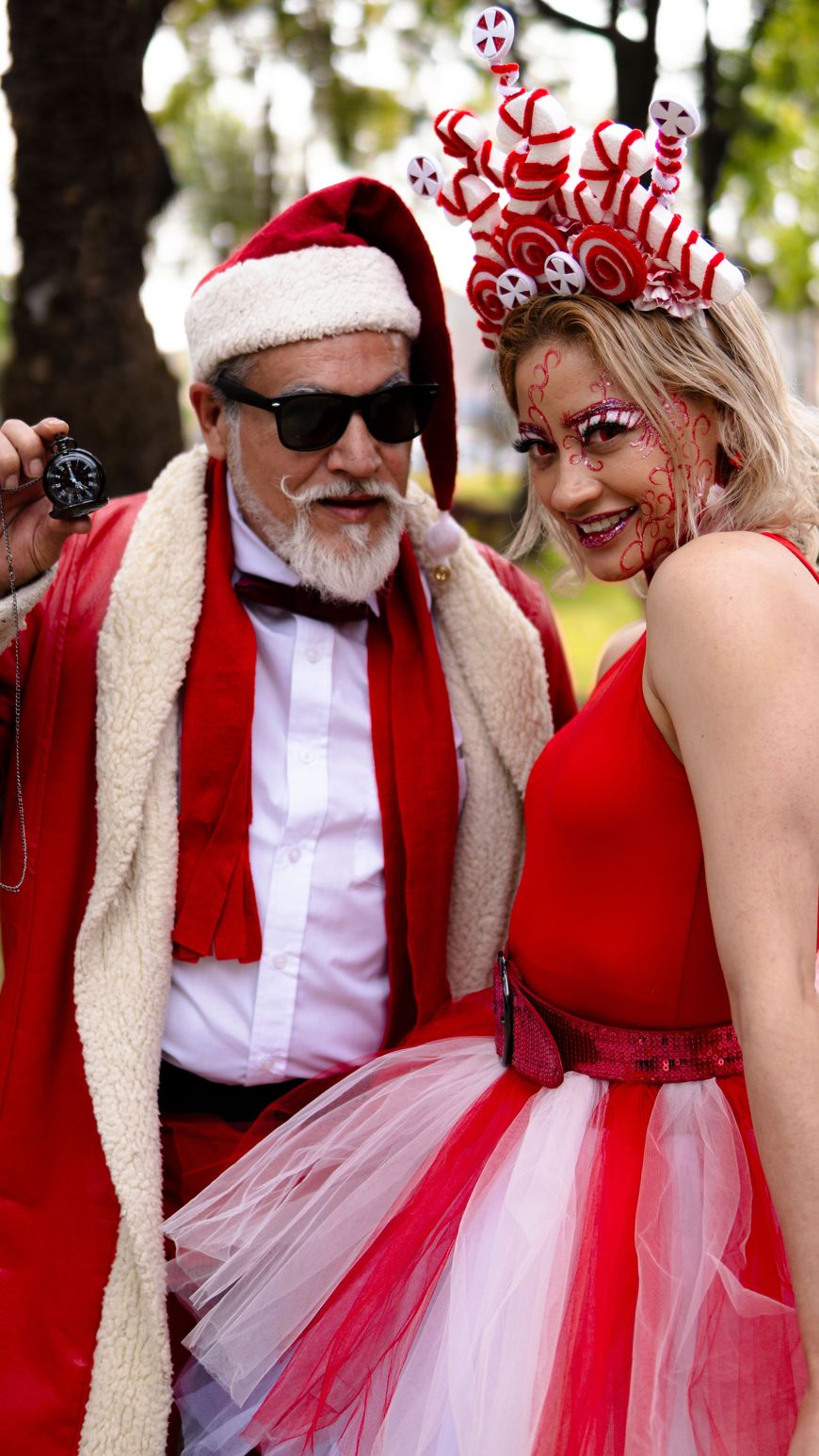 A couple in matching red costumes, one dressed as Santa with a red robe and sunglasses, the other wearing a candy cane themed headpiece and red tulle skirt.