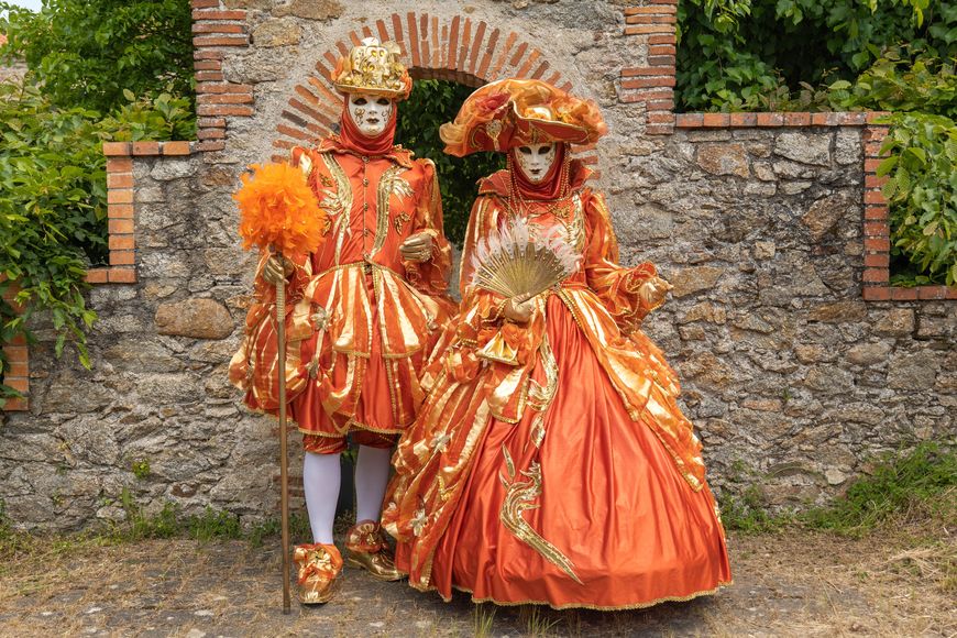 A couple in elaborate orange and gold Venetian carnival costumes with full face masks, standing by a stone archway.