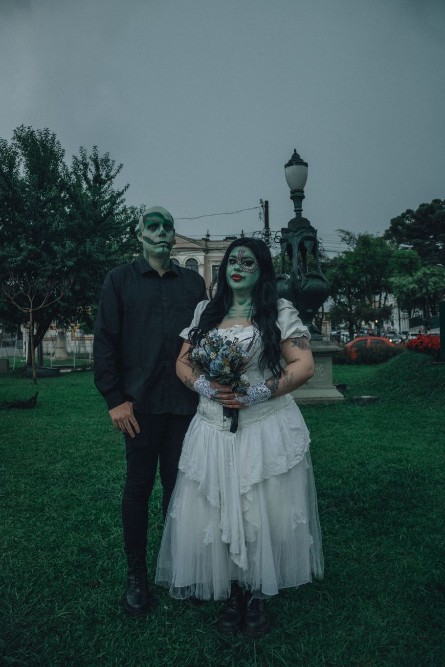 A couple in zombie wedding costumes with green face paint, one in a black suit and the other in a torn white wedding dress holding a bouquet.