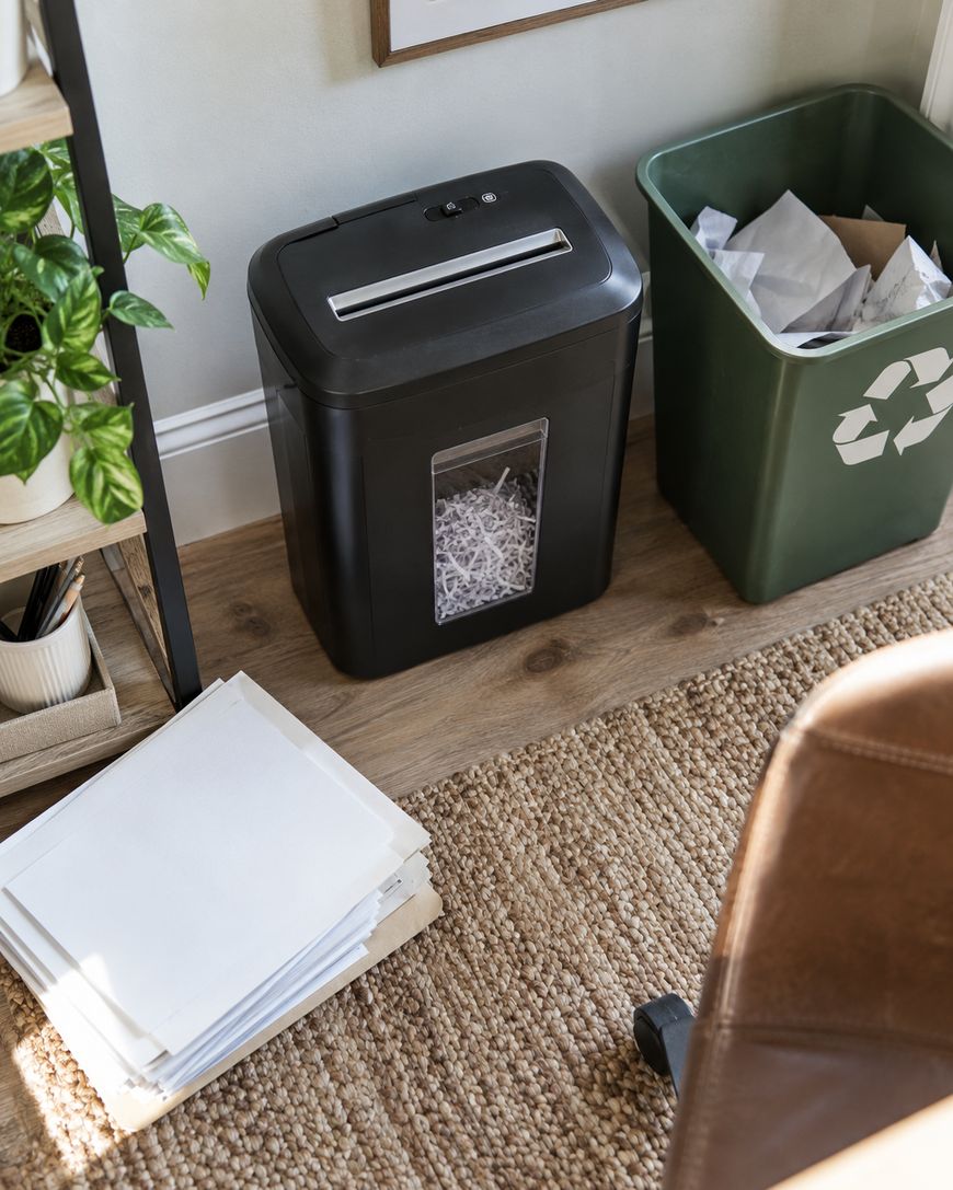 A paper shredder sits next to a green recycling bin filled with paper, a stack of unused paper on the floor, a plant on a shelf, and part of a brown chair on a woven rug.