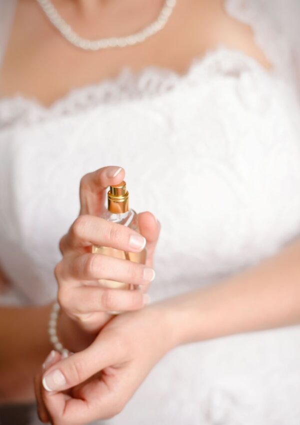 A bride in a white lace dress applies perfume to her wrist, wearing a pearl necklace and bracelet. Only her hands, jewelry, and upper torso are visible.