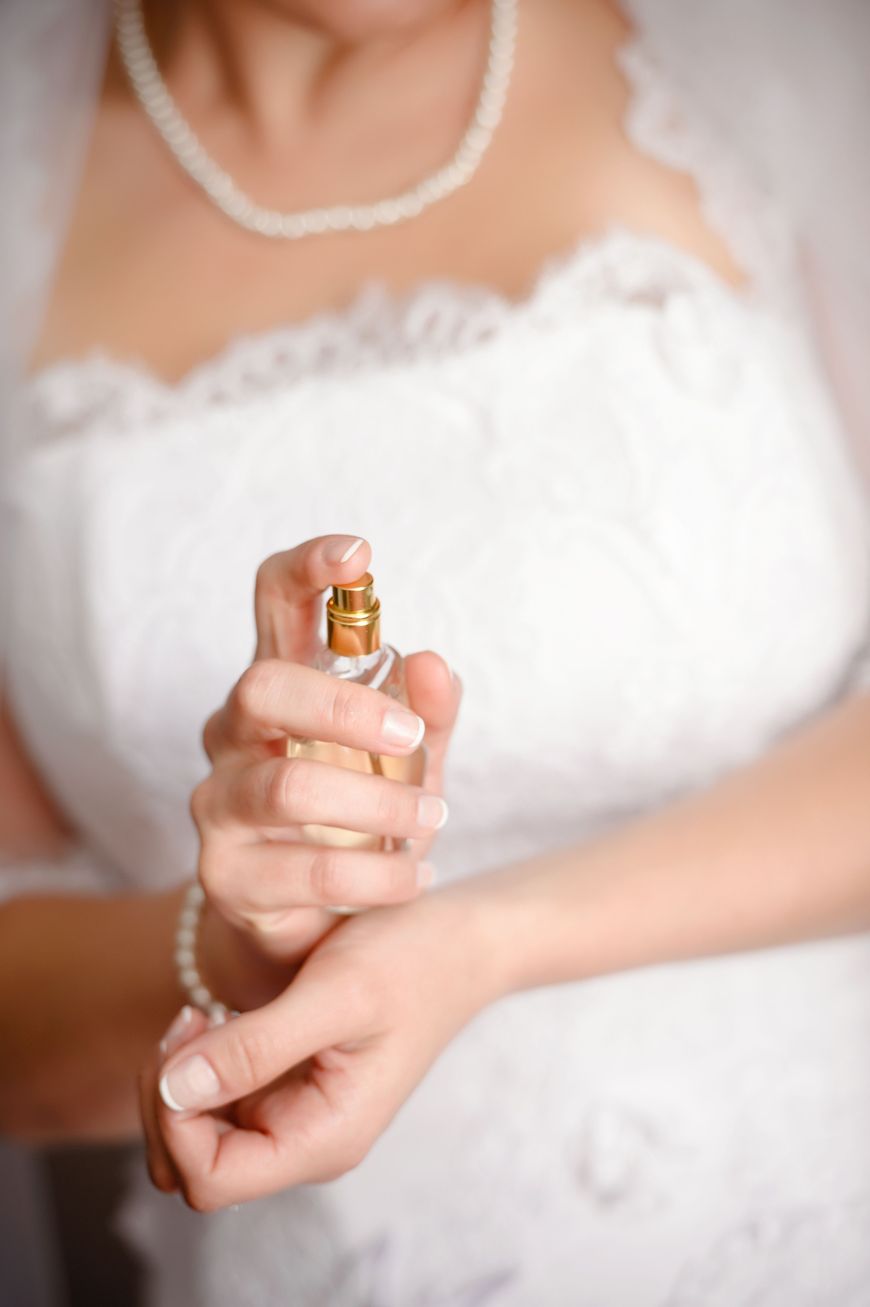 A woman in a white lace dress sprays perfume on her wrist. She wears a pearl necklace and bracelet, with soft focus on her upper body and hands.