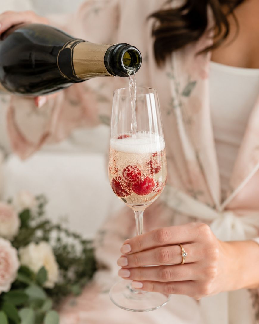 A person pours sparkling wine into a flute glass with raspberries. The person wears a light floral robe and has a ring on their finger. Flowers are blurred in the background.